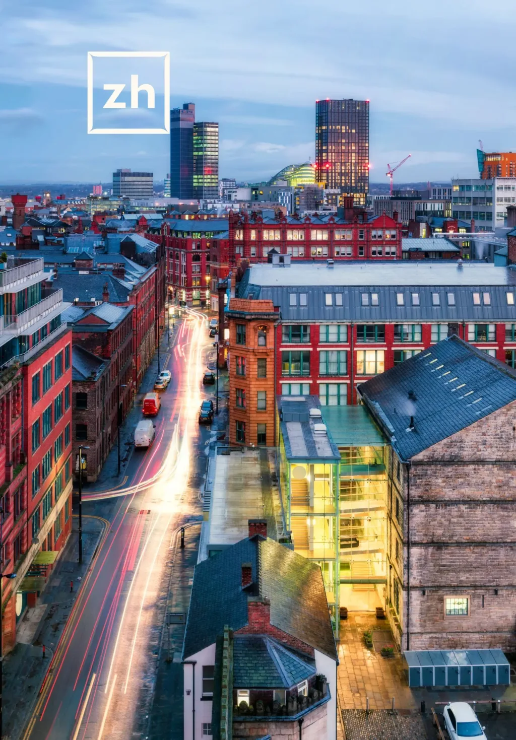 City skyline at dusk with light trails from traffic on a street lined with colorful buildings, tall towers in the background, and a construction crane visible. A 